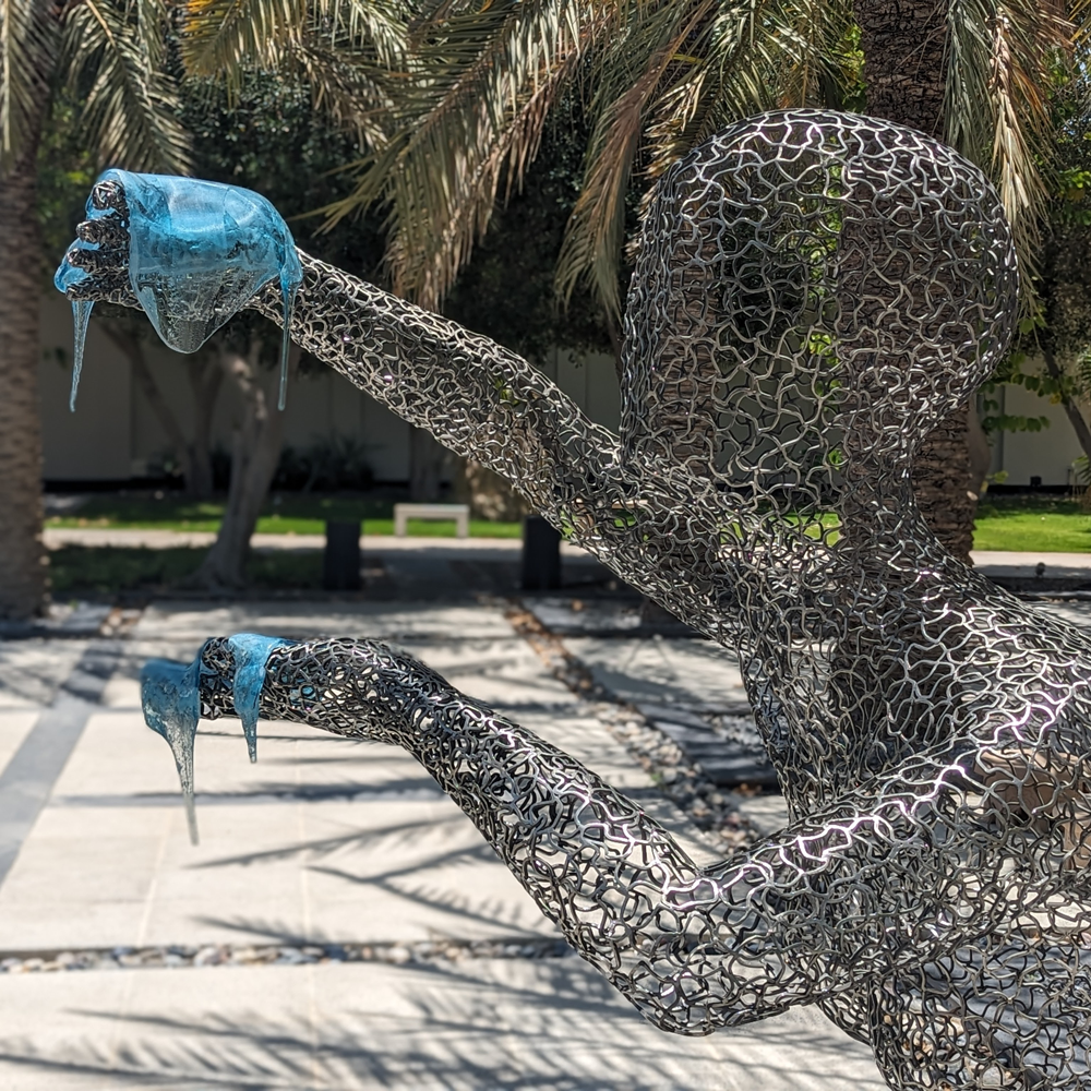 Stainless steel figure sculpture with blue glass elements, displayed outdoors